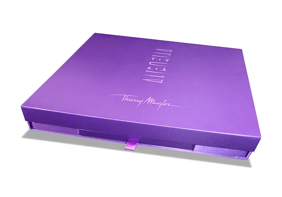 Coffret Thierry Mugler avec couvercle à rabat aimanté contrecollé de toile satin – impression à chaud ton sur ton – grille de calage « trottoir » en microcannelure contrecollée de papier pelliculé – tiroir avec cale en carton gris contrecollé de papier brillant pelliculé – rubans extracteur et limiteurs d’ouverture d’angle – Classeur contrecollé rembordé de papier or avec marquage or et intercalaires en polypropylène violet sérigraphiés – découpe d’onglets.
